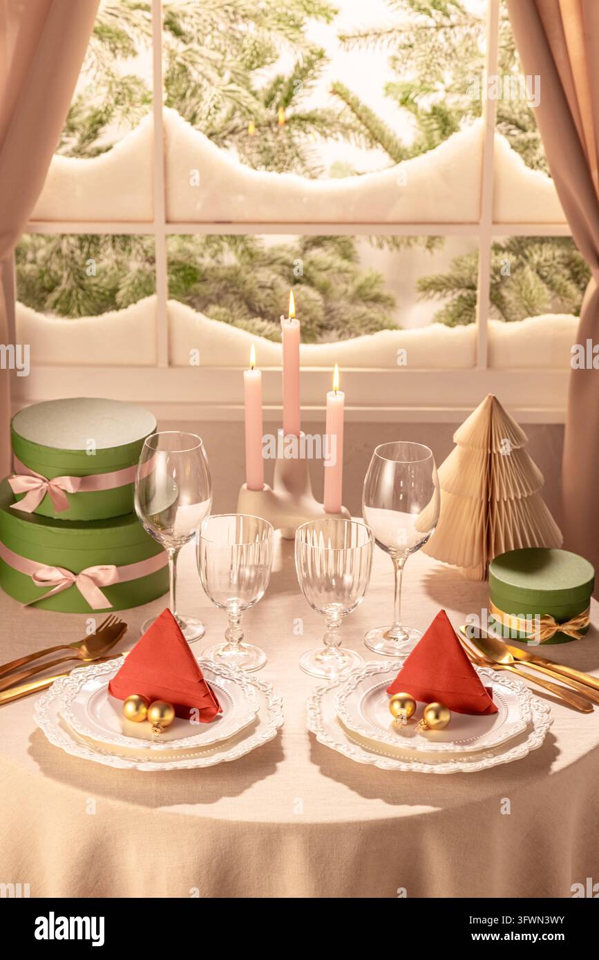 Vertikale Aufnahme eines symmetrisch gedeckten Weihnachtstisches mit roten Baerservietten, goldenen Weihnachtskugeln, leuchtenden rosa Kerzen, Papierbaum und Grün Stockfoto