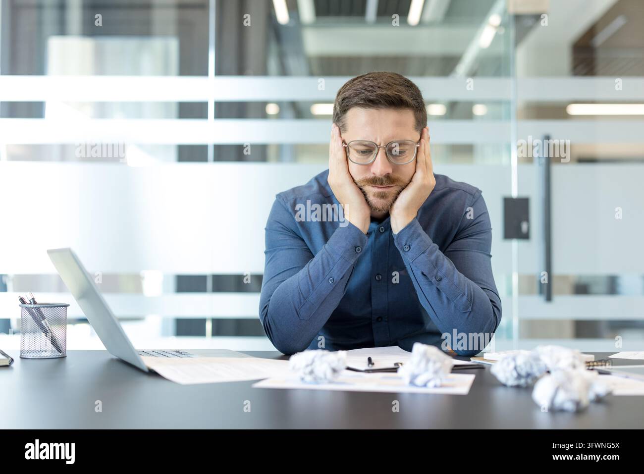 Frustrierter Büroangestellter sitzt an einem modernen Schreibtisch neben einem Laptop und verstreuten Dokumenten, hält den Kopf mit beiden Händen und steht allein vor Arbeitsdruck, Fehlern, Stress und knappen Fristen. Stockfoto