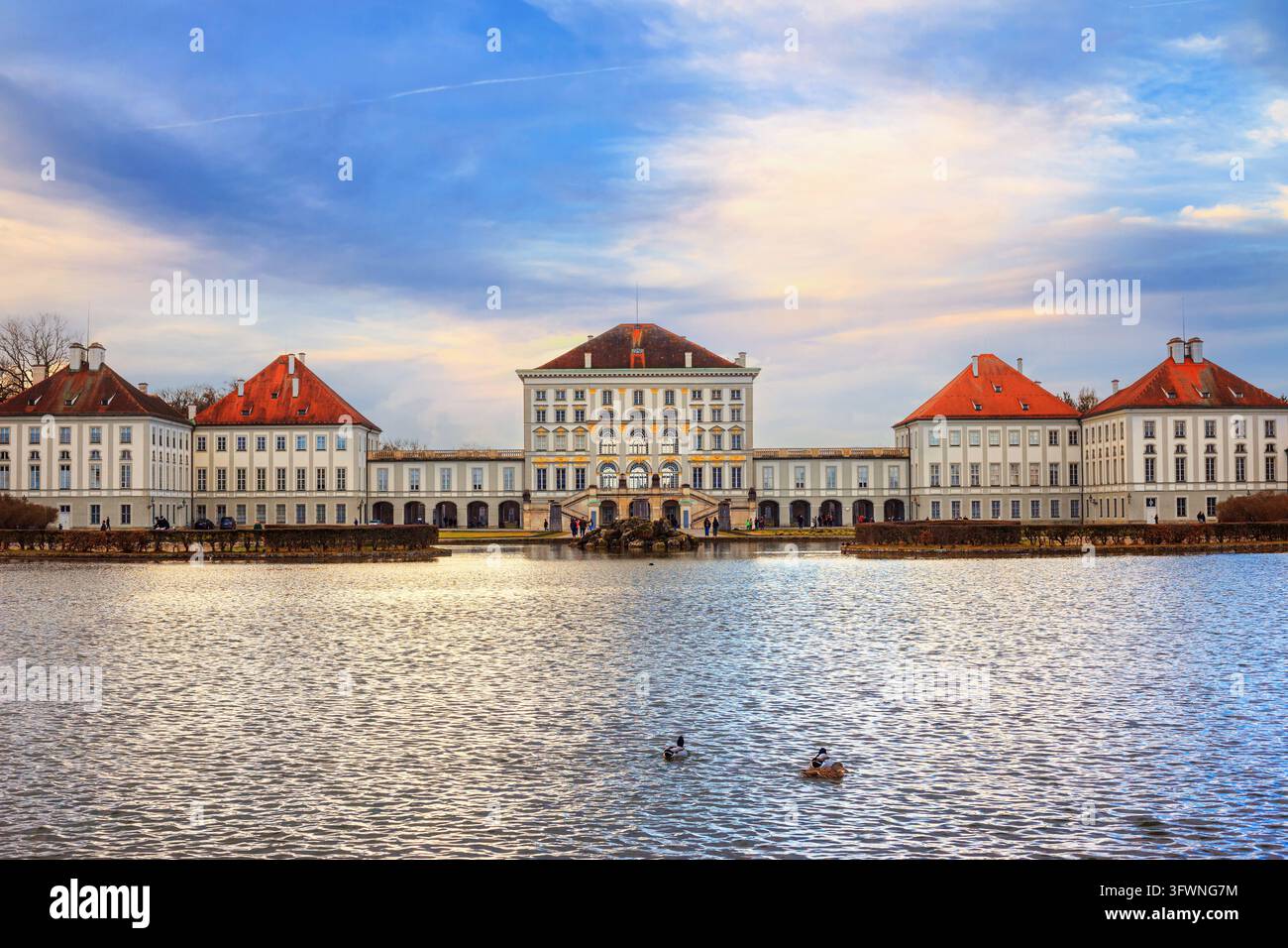 Ein weitwinkliges, dramatisches Augenhöhe-Foto des großen Schlosses Nymphenburg in München, Deutschland, über einen ruhigen See während der späten Goldenen Stunde Stockfoto
