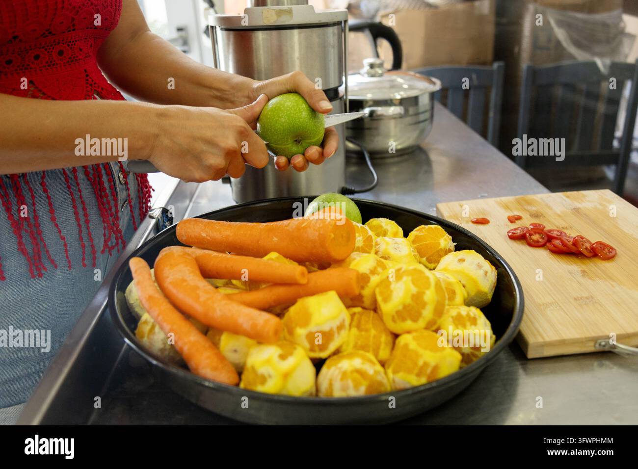 Nahaufnahme einer Frau, die einen grünen Apfel neben Karotten und geschälter Zitrusfrucht in einer Küche schält und frische Zutaten für hausgemachten Saft zubereitet. Stockfoto