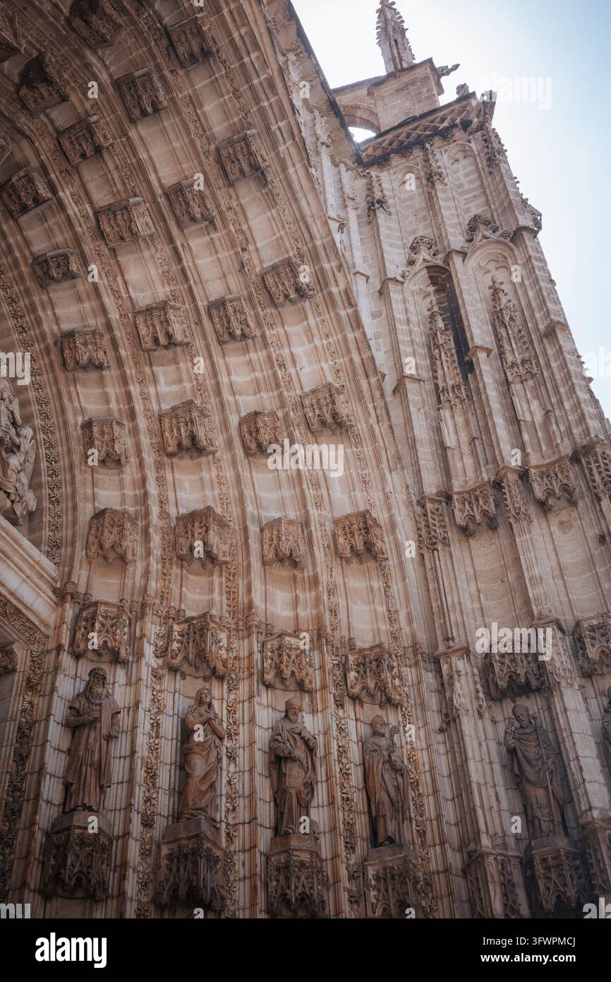 Niedrig gewinkelte Ansicht einer verzierten gotischen Kathedralenfassade in Sevilla, Spanien, mit geschnitzten steinernen Heiligen, spitzen Bögen, aufwendigem Maßwerk, Säulen und h Stockfoto