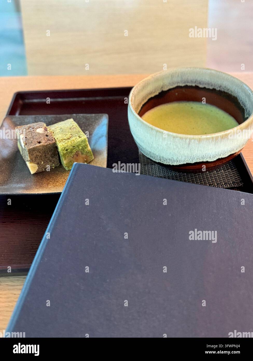 Schale mit Matcha-Grüntee, serviert mit traditionellen japanischen Desserts auf einem schwarzen Tablett, das auf einem Holztisch in einer einfachen und ruhigen Teeraum-Atmosphäre steht Stockfoto
