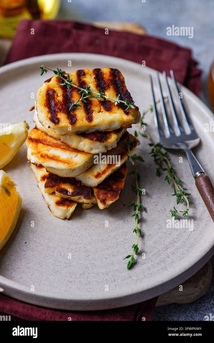 Goldener gegrillter griechischer Halloumi-Käse, gestapelt auf einem Keramikteller mit frischem Thymian, Zitronenspaltern und Gabel, wodurch eine rustikale mediterrane Vorspeise entsteht Stockfoto