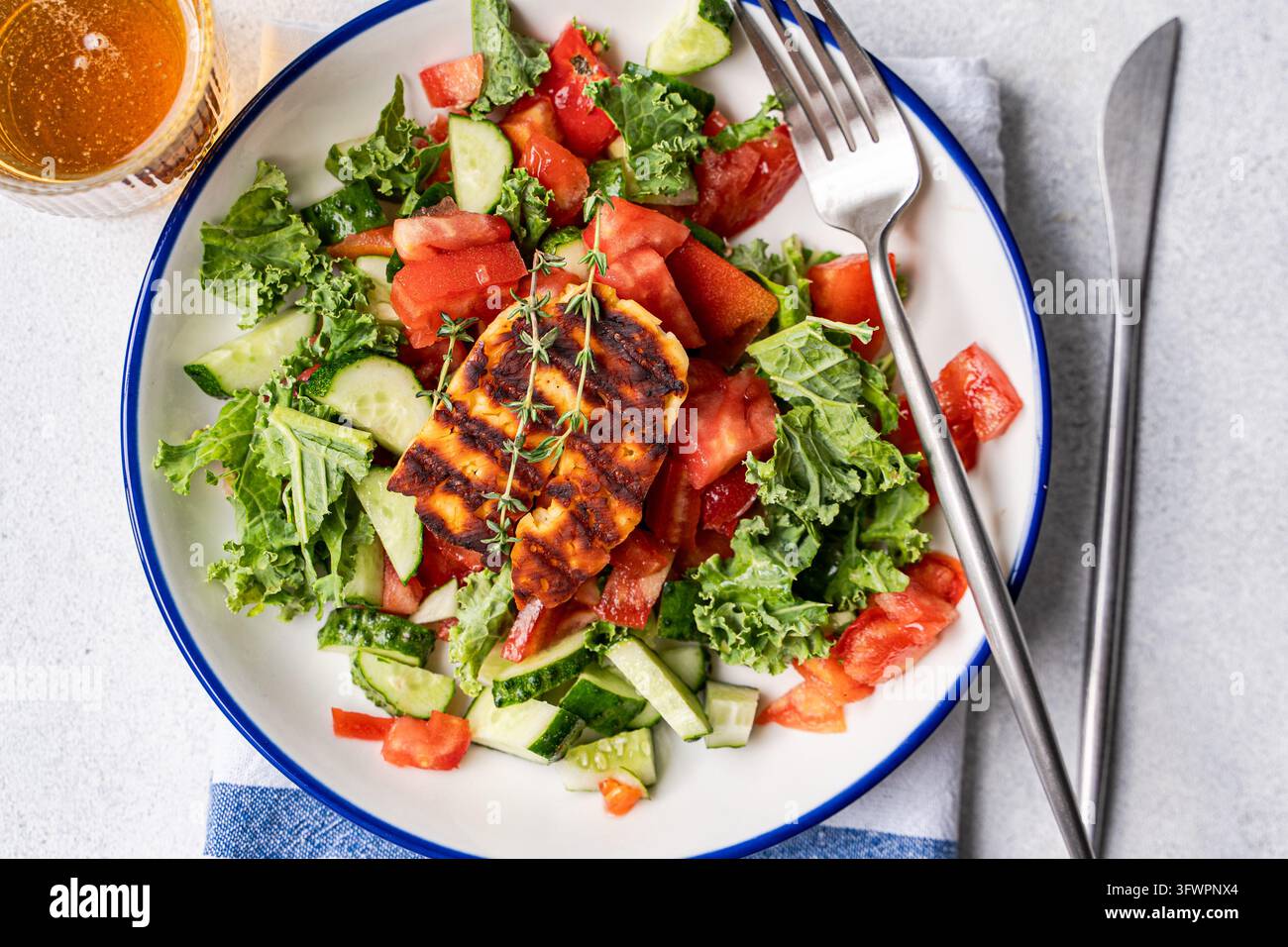 Aufsichtsansicht von griechischem Halloumi-Salat mit gegrilltem Käse, Kohl, Gurke, Tomate und frischem Thymian, serviert auf einem Teller mit Gabel, Messer und kaltem Bier. Stockfoto