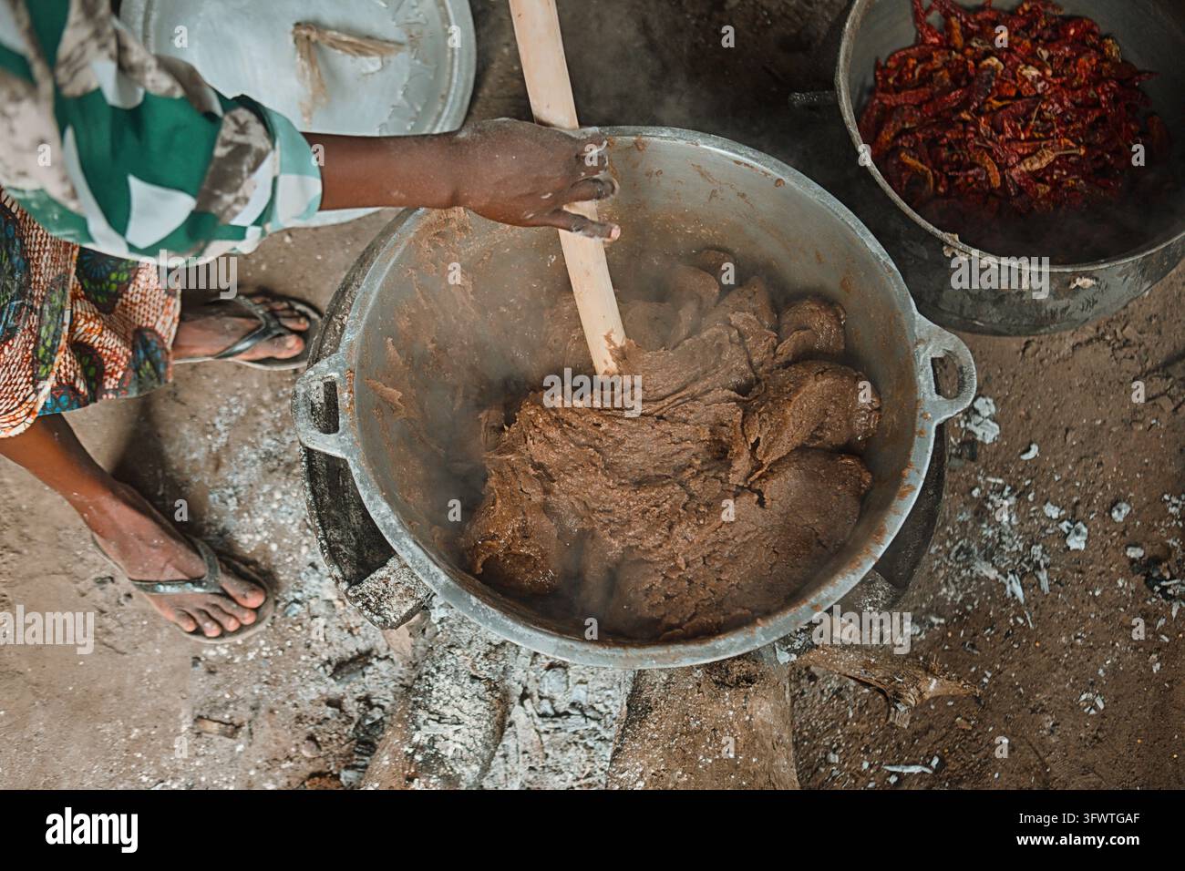 Ansicht einer Person, die einen großen Topf mit traditionellem Essen über einem offenen Feuer rührt, mit einer Schüssel getrockneter Peperoni daneben in Igbo-Ora, Oyo, Nigeria. Stockfoto