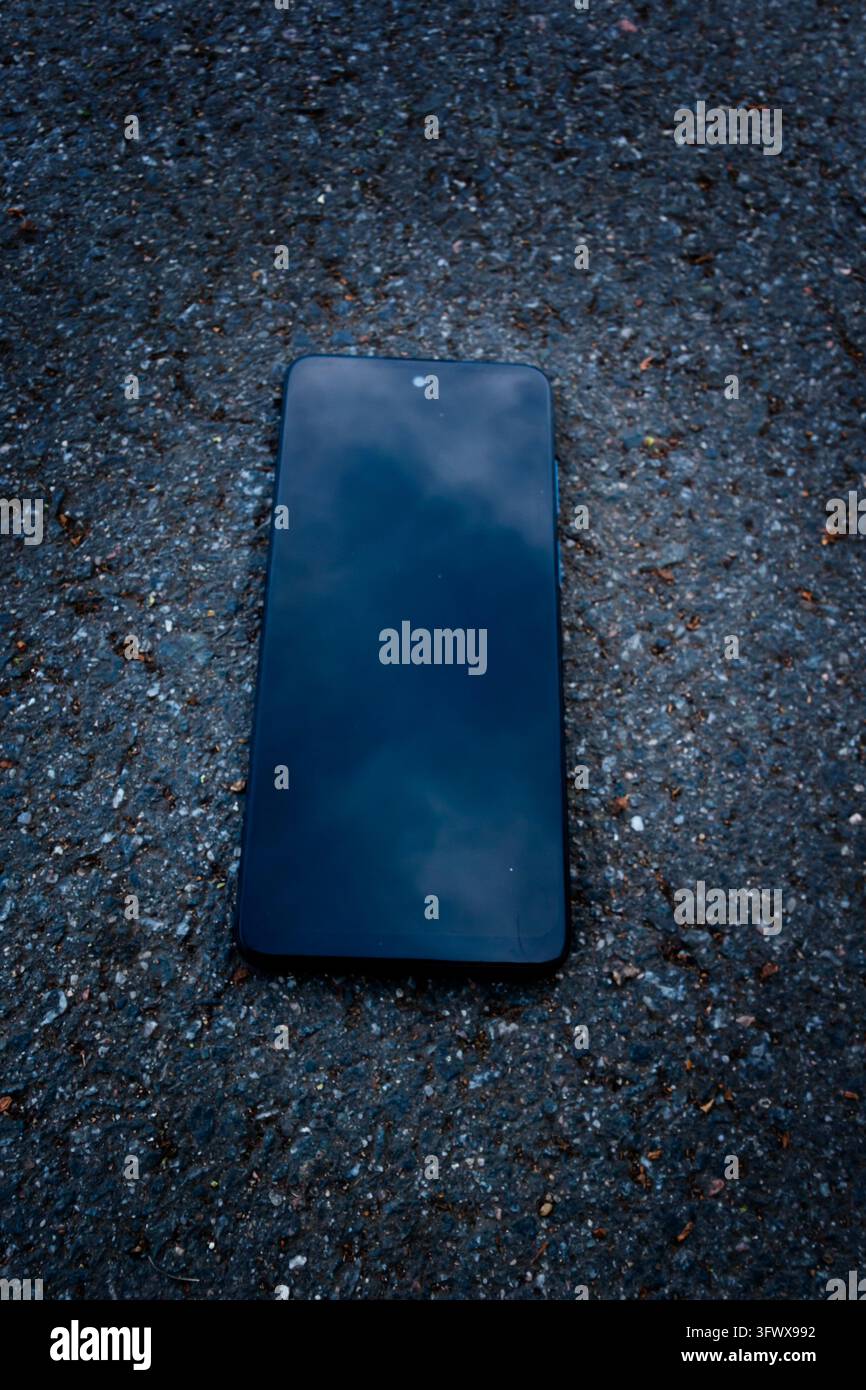 Schwarzes Smartphone liegt mit der Vorderseite nach oben auf rauem Asphalt, sein intaktes dunkles Display spiegelt einen bewölkten Himmel in einer top-down-Außenaufnahme wider Stockfoto