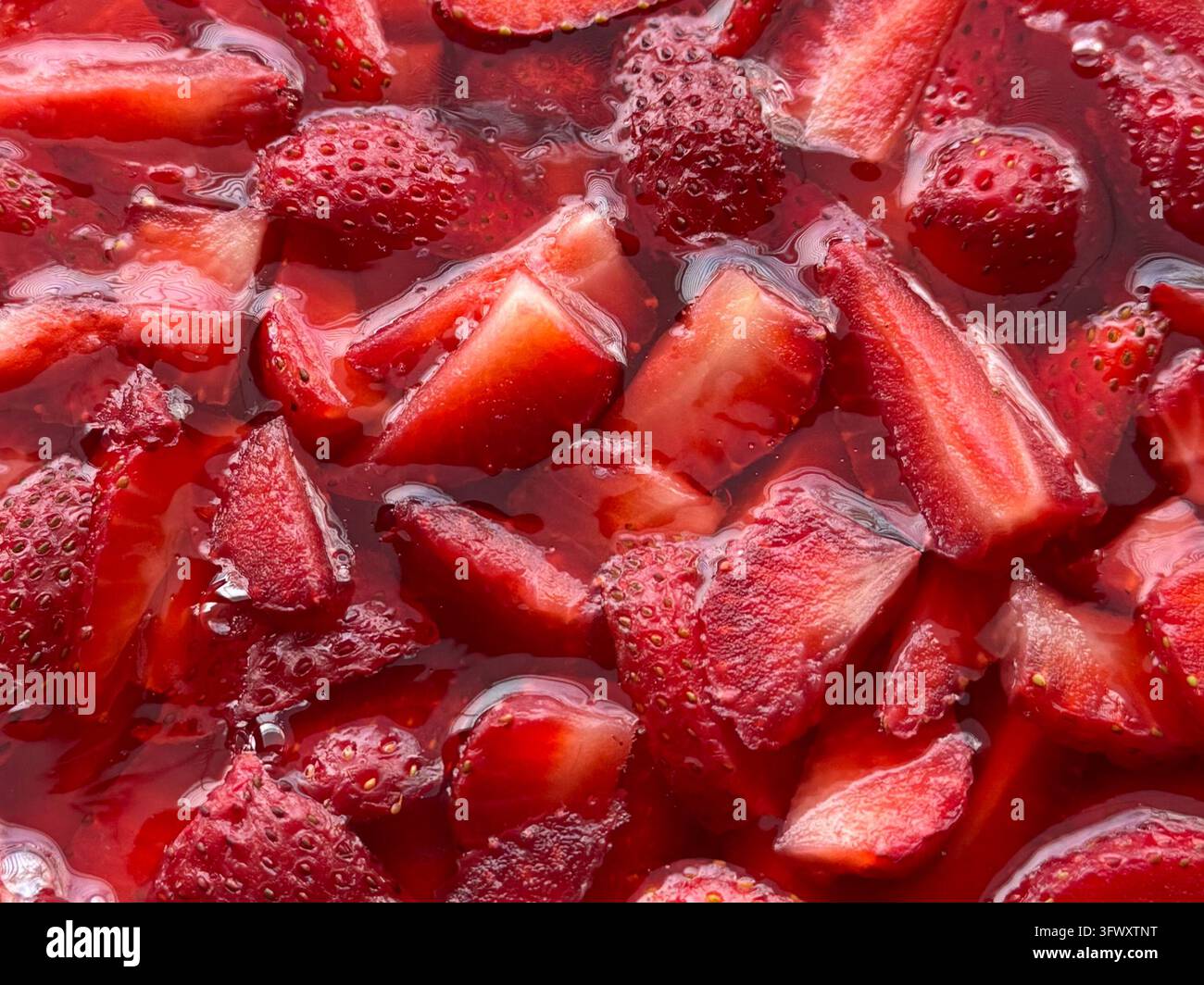Vollformat-Nahaufnahme von halbierten und viertelten reifen Erdbeeren, die in ihrem eigenen roten Saft und Sirup glänzen. Die glänzende Frucht zeigt Samen und saftiges Fleisch - Smartphone-aufgenommenes Stockfoto