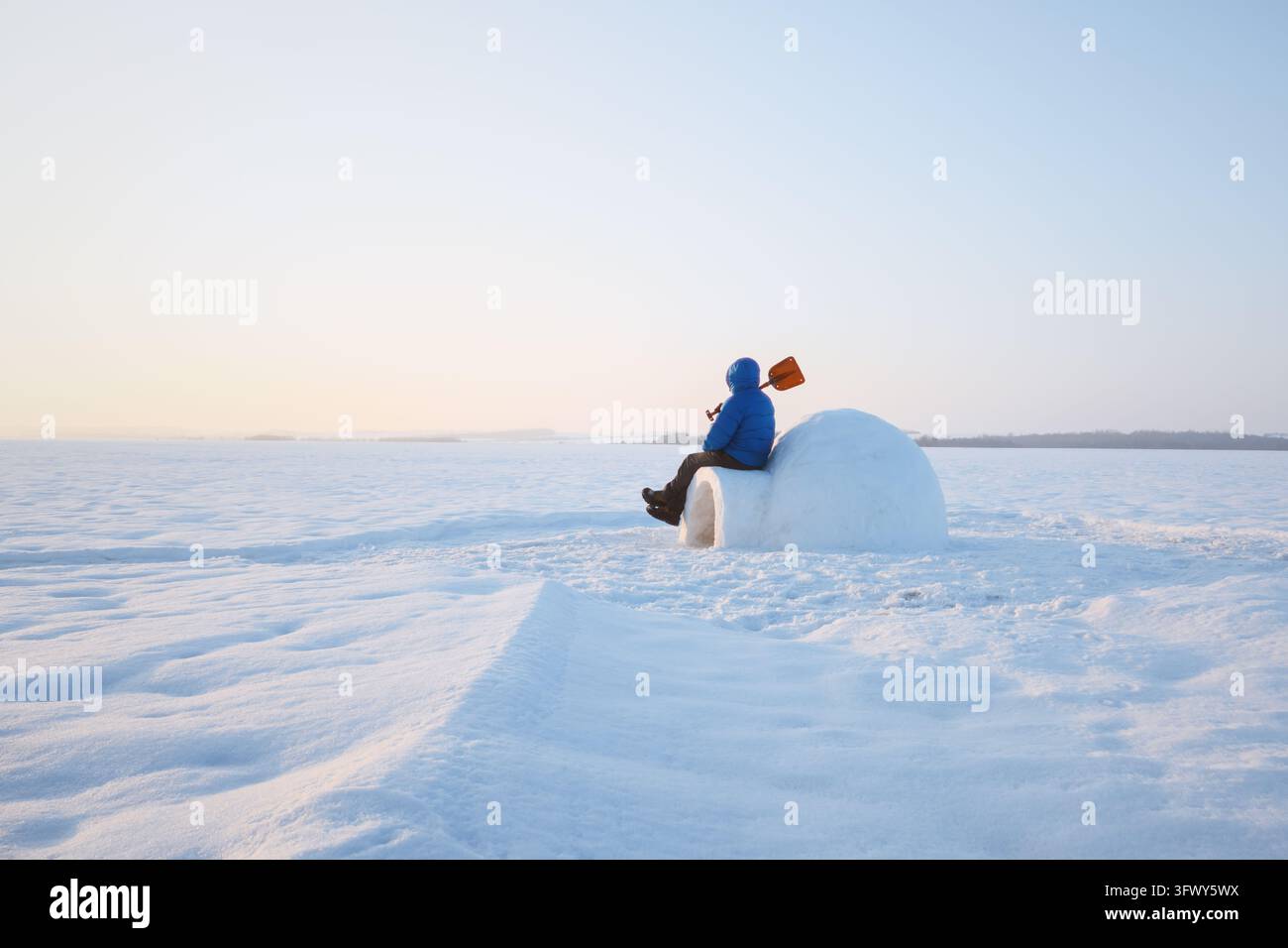 Mann in blauer Winterkleidung sitzt neben einem Schnee-Iglu in einer weiten gefrorenen Landschaft. Blauer Himmel und unberührter Schnee schaffen eine friedliche arktische Campingszene Stockfoto
