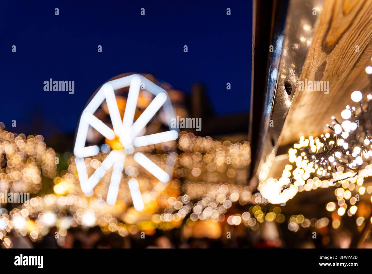 Unscharfer Weihnachtsmarkt leuchtet mit warmen Lichterketten und beleuchtetem Riesenrad unter tiefblauem Abendhimmel. Weiches goldenes Bokeh erzeugt festliche Stimmung Stockfoto