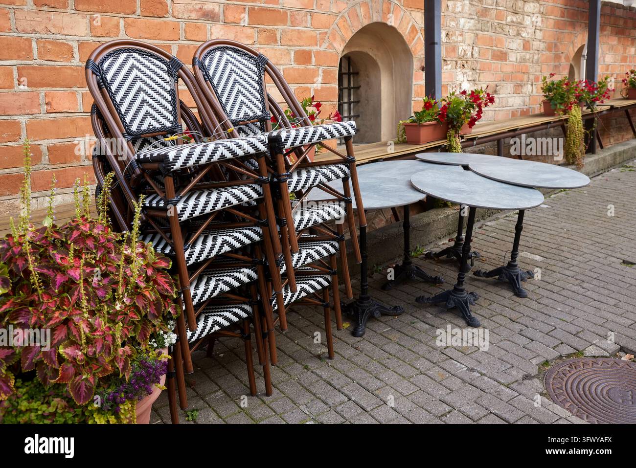 Saisonende Café-Schließung mit gestapelten Außenstühlen, Bistrotischen und Blumenkästen neben einem Backsteingebäude. Saisonale Restaurantschließung Stockfoto