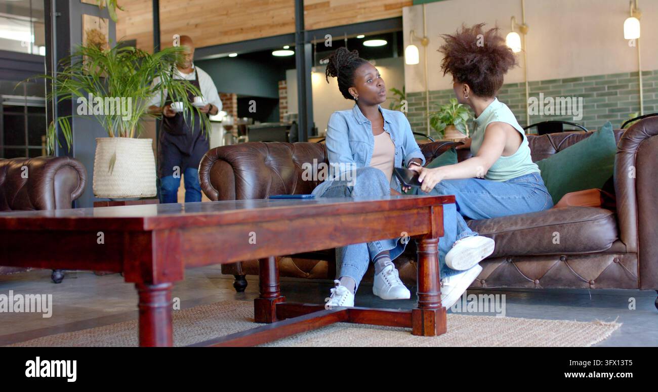 Afrikanische Freundinnen, die auf einem Ledersofa in einem Café sitzen und sich unterhalten, mit Smartphone und Handtasche Stockfoto