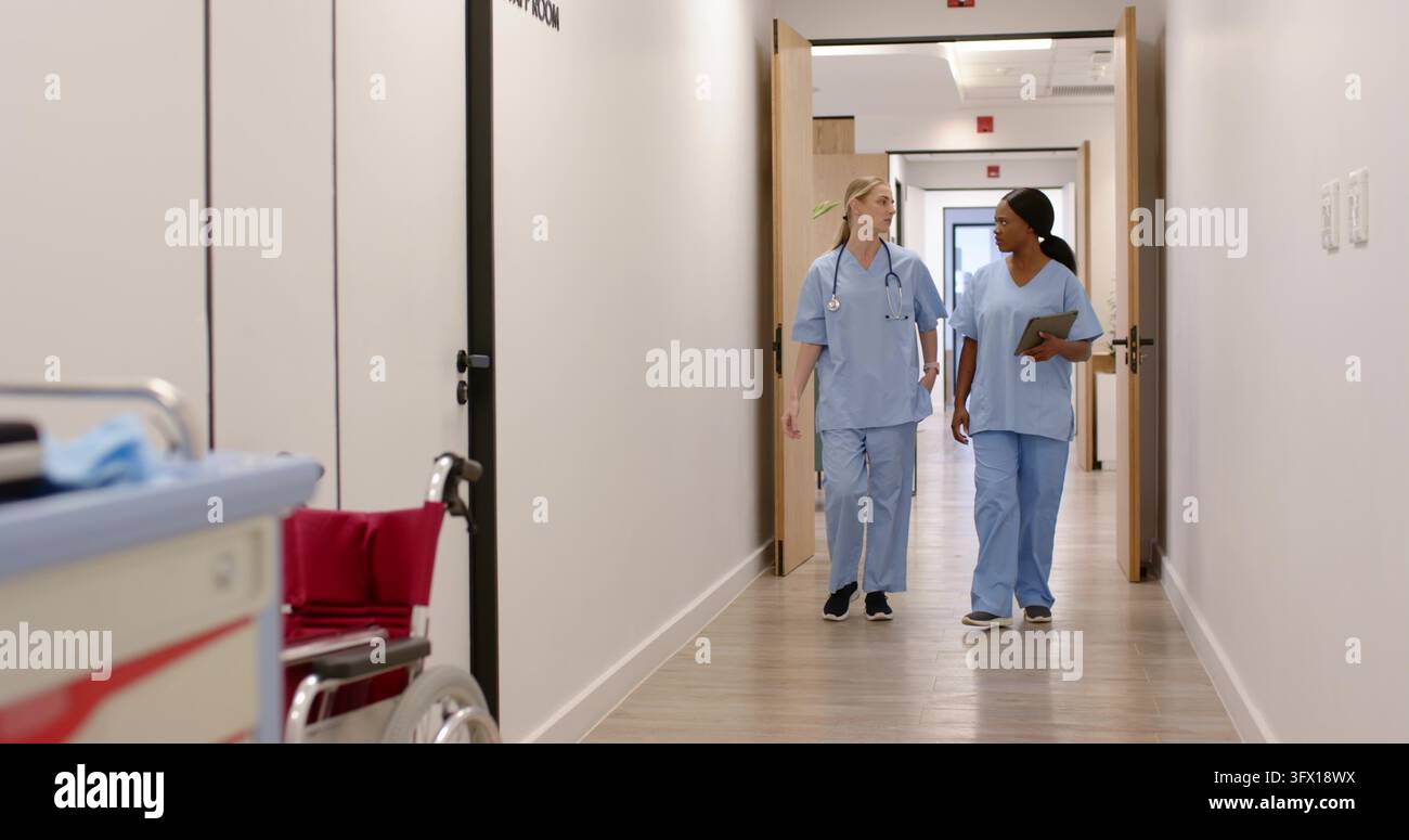 Verschiedene Krankenschwestern gehen und unterhalten sich in blauen Kitteln den Krankenhausflur entlang, Stethoskop-Tablet Stockfoto