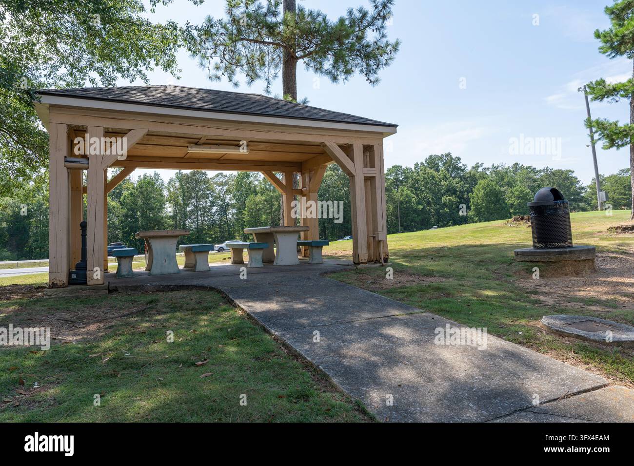 Holzschild mit Betonpicknicktischen und Bänken an einem ruhigen Rastplatz am Straßenrand, umgeben von grünem Gras und hohen Bäumen. Überdachte Außenseite Stockfoto