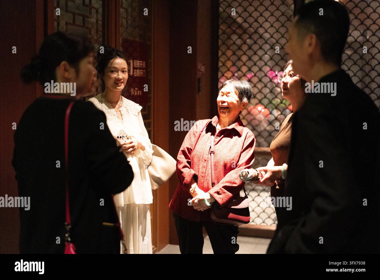 Eine fröhliche multigenerationale Gruppe asiatischer Frauen, die nachts drinnen lachen und sich unterhalten, mit traditionellen chinesischen Architekturelementen, darunter ein fischschuppenmusteriger Schirm und ein Holzrahmen mit roter Beschriftung in chinesischen Schriftzeichen, warme Umgebungsbeleuchtung, die ausdrucksstarke Gesichter und kulturelle Kleidung hervorhebt Stockfoto