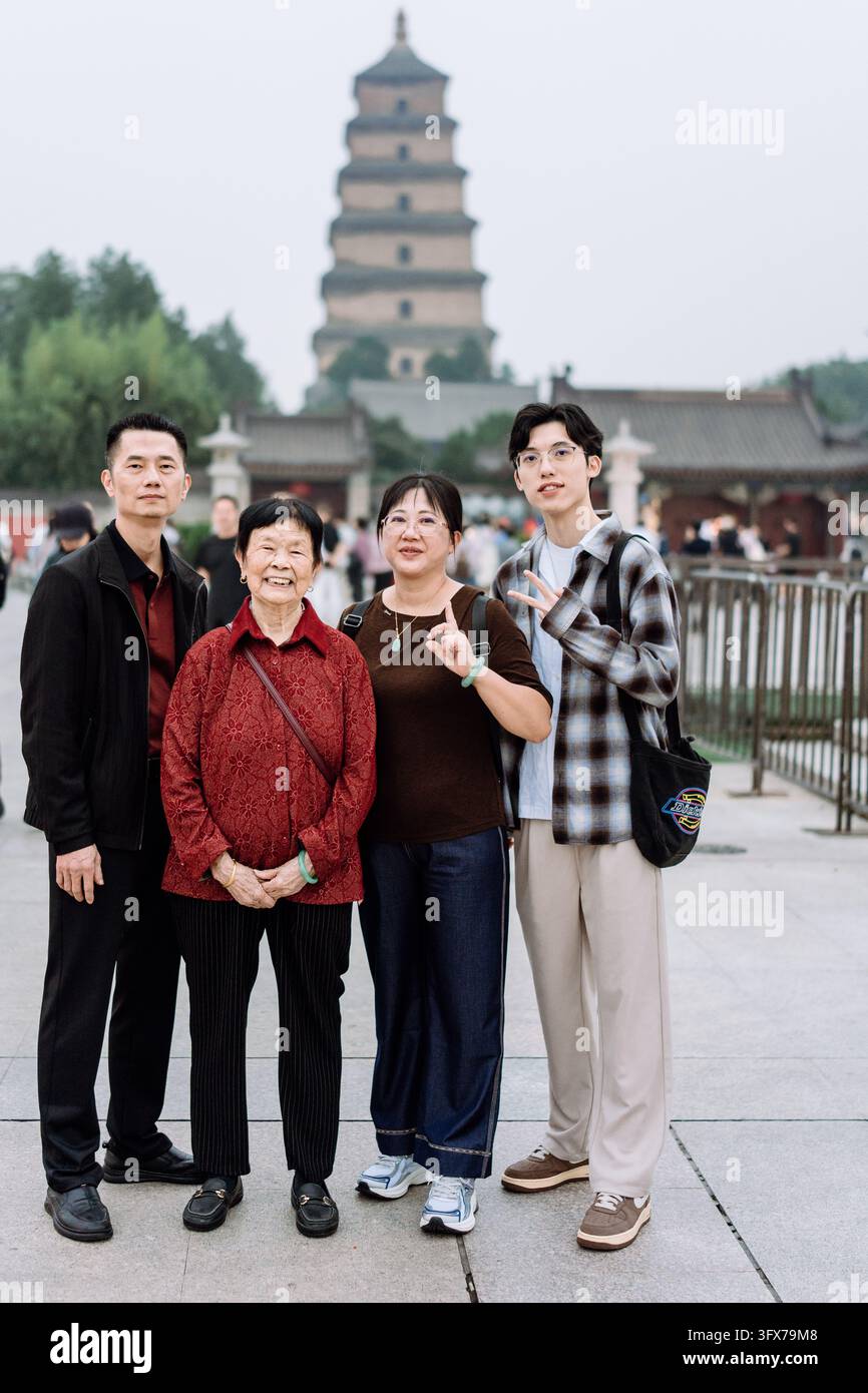 Eine multigenerationale chinesische Familie, die an einem bewölkten Tag vor der Großen Wildgans-Pagode in Xi’an, Provinz Shaanxi, China, für ein Foto posiert; die ältere Frau trägt eine traditionelle rot bestickte Jacke, während jüngere Mitglieder lässige moderne Kleidung tragen, und fängt einen Moment kultureller Kontinuität und Tourismus an diesem historischen buddhistischen Wahrzeichen ein. Stockfoto