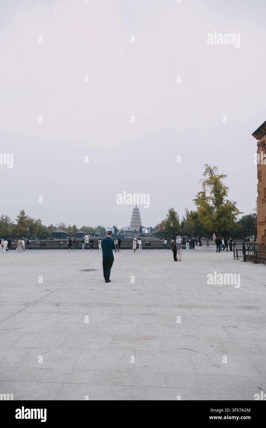 Breitwinkelaufnahme des Großen Wildgans-Pagoden in Xi’an, China, der am Ende eines weitläufigen gepflasterten Platzes unter einem bewölkten Himmel steht, mit vereinzelten Touristen, die herumgehen und beobachten, flankiert von herbstlichen Bäumen und traditionell stilisierten Gebäuden auf der rechten Seite Stockfoto