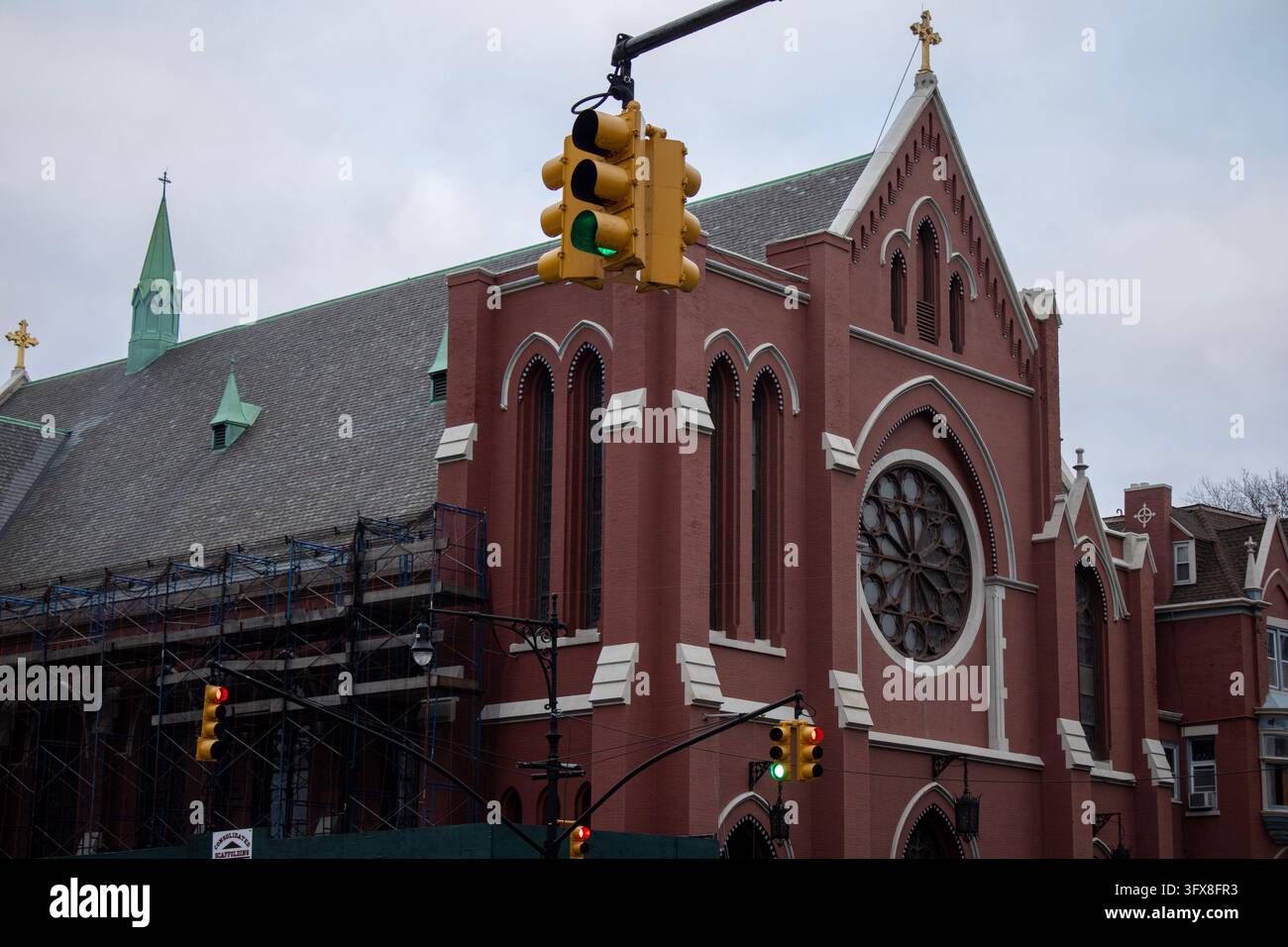 New York City, USA – 31. Januar 2024: Rote Backstein-Gotikrevival-Kirche mit einem Rosettenfenster und einem Turm neben einer Ampel an einem bewölkten Tag. Stockfoto