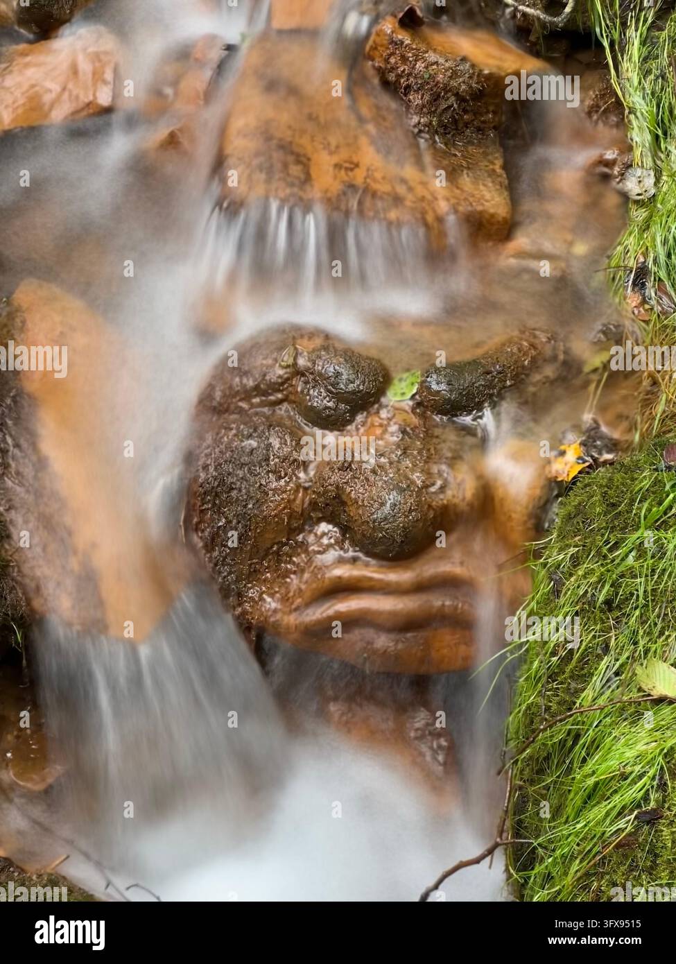 Steingesichtsskulptur, in feuchte Felsen gehauen, unter fließendem Wasser aus einem seidigen Wasserfall. Wasser fließt sanft über einen braunen Stein, der zu einem Gesicht gemeißelt ist. - Smartphone-aufgenommenes Stockfoto