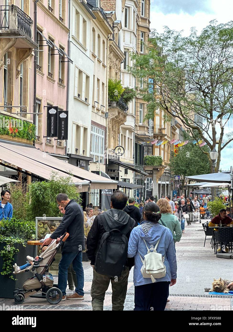 Menschen schlendern eine belebte europäische Fußgängerzone entlang mit Straßencafés und historischen Gebäuden. Fußgänger schlendern eine gepflasterte Stadtstraße entlang - Smartphone-aufgenommenes Stockfoto