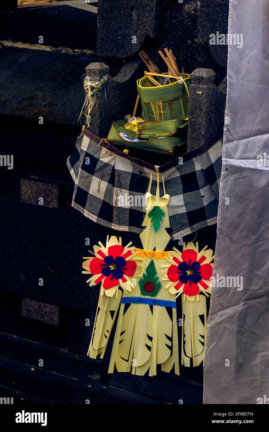 Nahaufnahme eines schwarz-weiß karierten Stoffes, der mit einem hellen handgefertigten Papierornament und Palmblättern geschmückt ist und an einer dunklen Tempelmauer hängt. Stockfoto