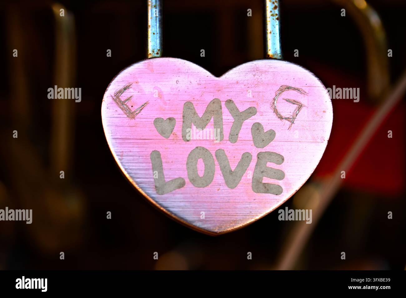 Ein verwittertes rosa herzförmiges Schild mit den Worten "MEINE LIEBE" hängt an Metallstäben und hebt sich von einem warmen, sanft verschwommenen Hintergrund ab. Stockfoto