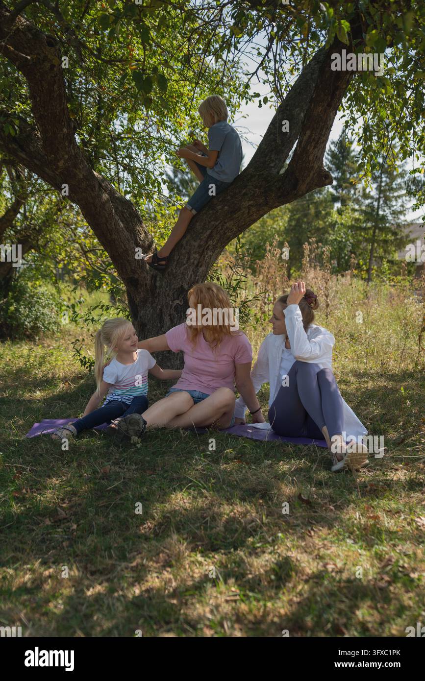 Mehrgenerationen-Familienmitglieder, die Zeit zusammen im Freien verbringen, ein Junge liest im Baum, während Frauen und ein Mädchen auf Matten auf dem Gras sitzen Stockfoto