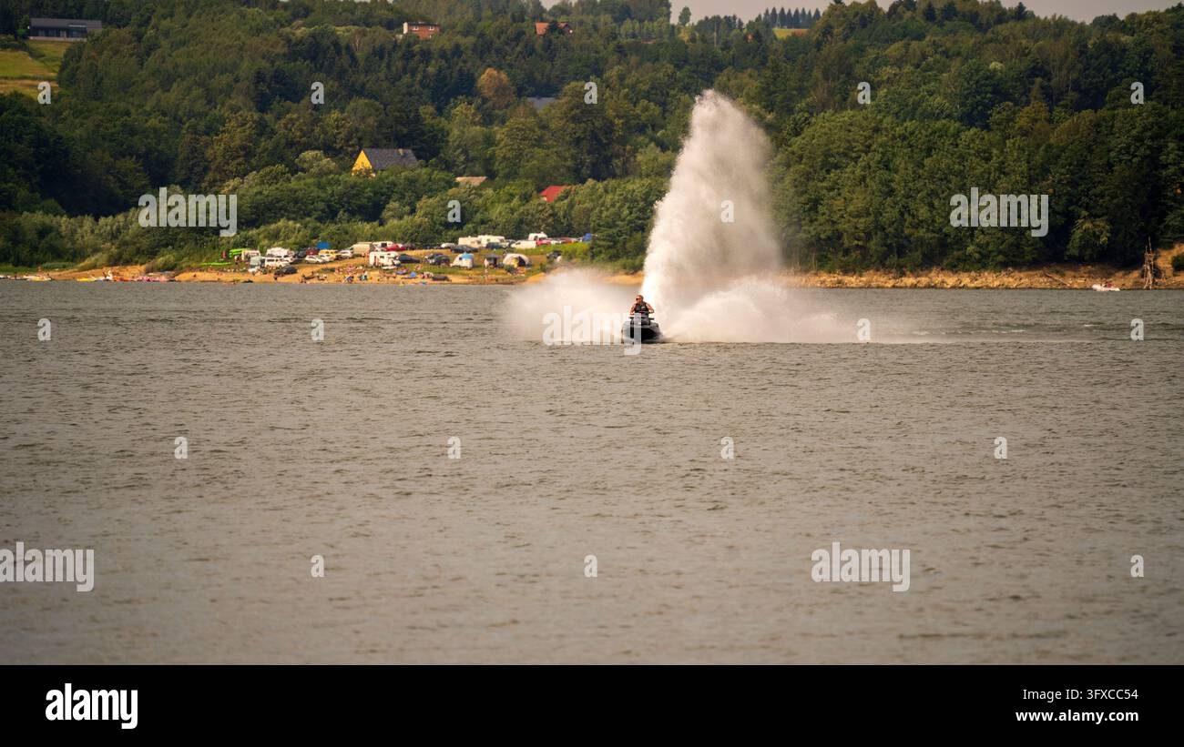 Jet-Ski-Fahrer erzeugt eine massive Wasserspritzfontäne, während er über den See rast, mit bewaldetem Ufer, Campingplatz und Häusern im Hintergrund an einem sonnigen Tag Stockfoto