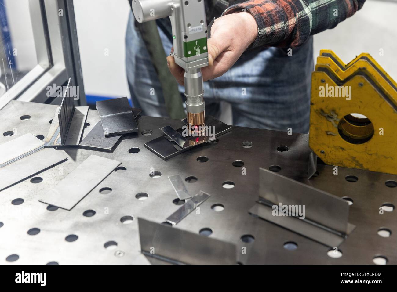 Bedienender Hände, die eine moderne handgeführte Laser-Schweißpistole halten, die Metallbleche auf einem Schweißtisch verbinden. Nahaufnahme von präziser Lasermetallbearbeitung Stockfoto