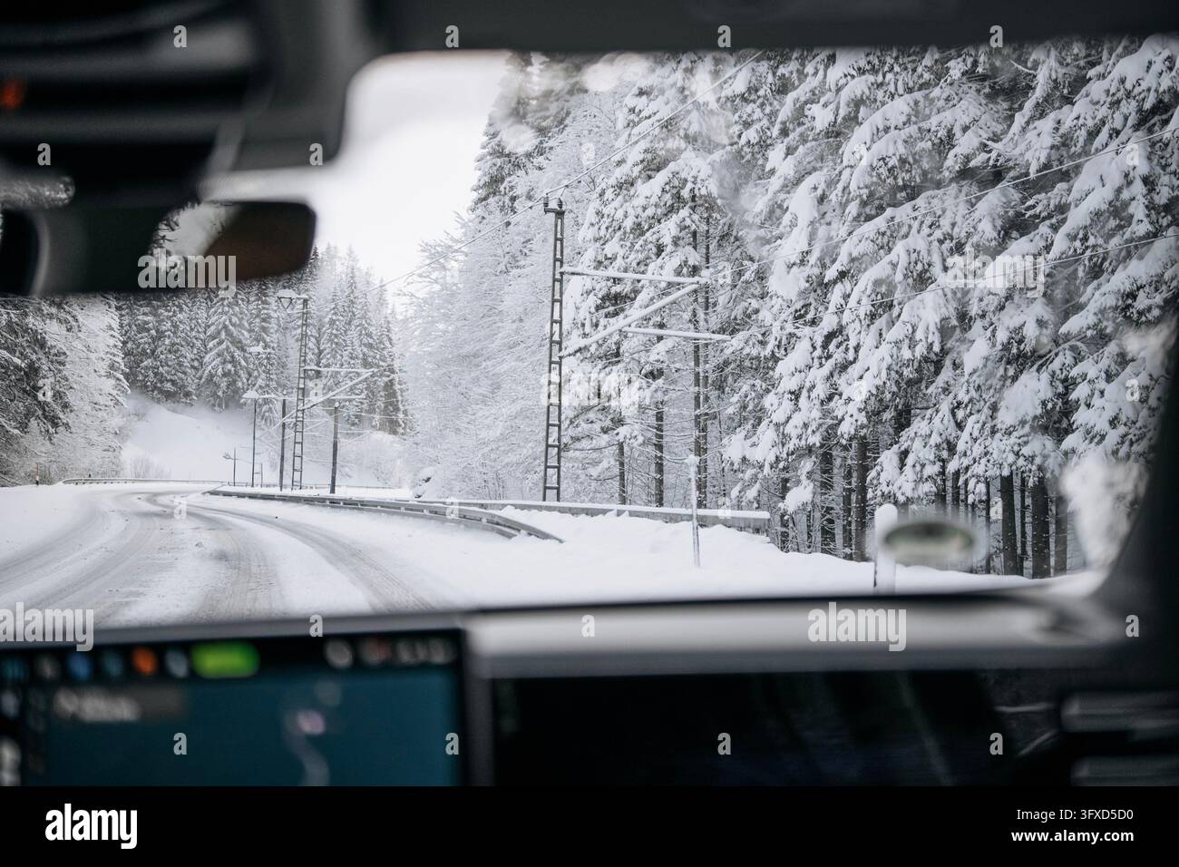 Blick aus einem Auto mit Lenkrad, Rückspiegel und Armaturenbrett im Bild, nach vorne entlang einer verschneiten Landstraße, die an alten Ti vorbeischwung Stockfoto