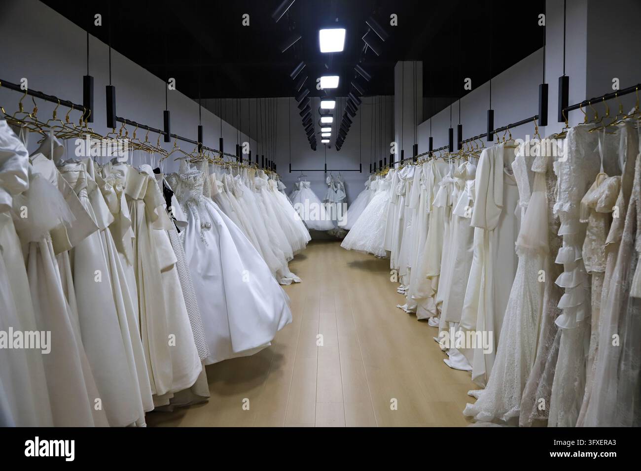 Innenraum eines Brautmodengeschäfts, an beiden Seiten hängen weiße Hochzeitskleider aus Spitze und Satin. Holzboden mit langen Deckenstrahlern schafft eine ele Stockfoto