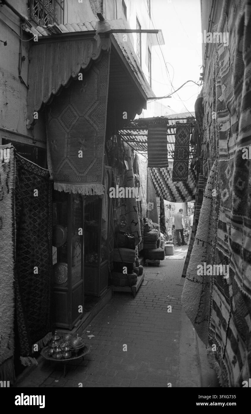 Schwarz-Weiß-Ansicht einer engen Marktgasse mit gemusterten Teppichen, die vor Geschäften hängen, und Metall-Teekannen auf einem Tablett. Stockfoto