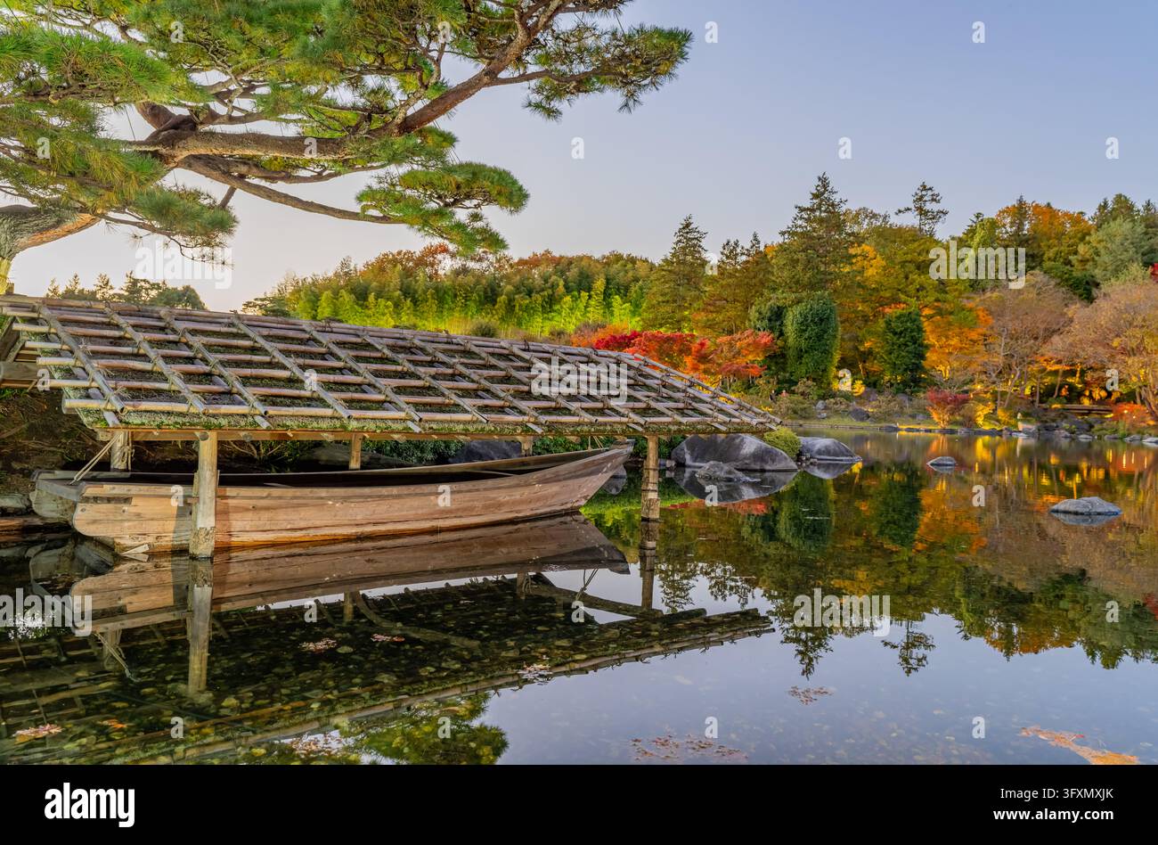 Ein traditionelles Holzboot mit einem Bambusdach ruht auf einem ruhigen Teich und spiegelt die umliegenden Bäume und das farbenfrohe Laub wider. Stockfoto