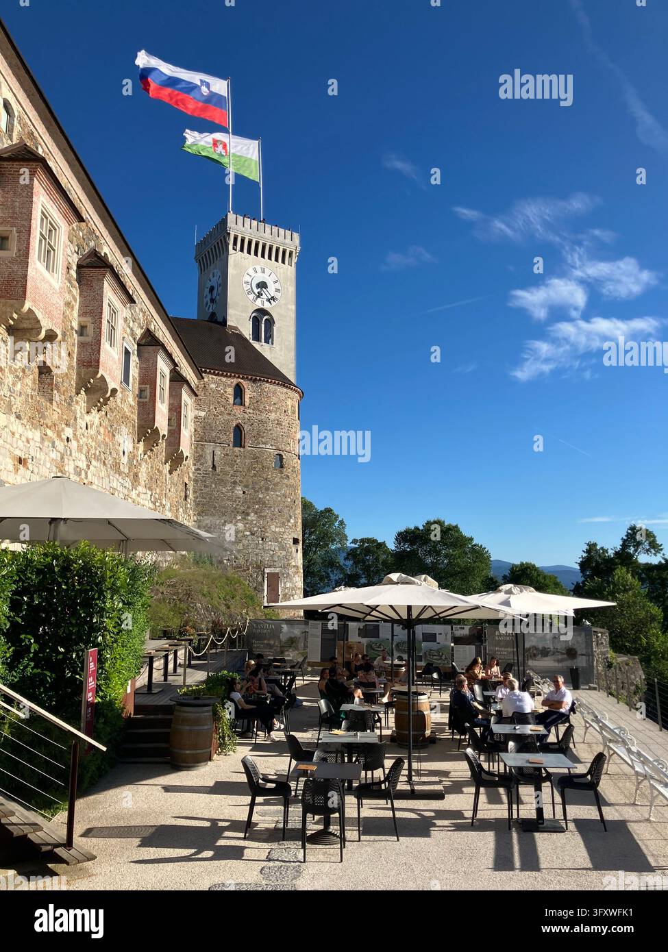 Besucher entspannen sich auf einer Außenterrasse eines Cafés auf der Burg von Ljubljana unter dem Glockenturm, der Fahnen in Ljubljana, Slowenien, an einem sonnigen Tag hisst. Stockfoto