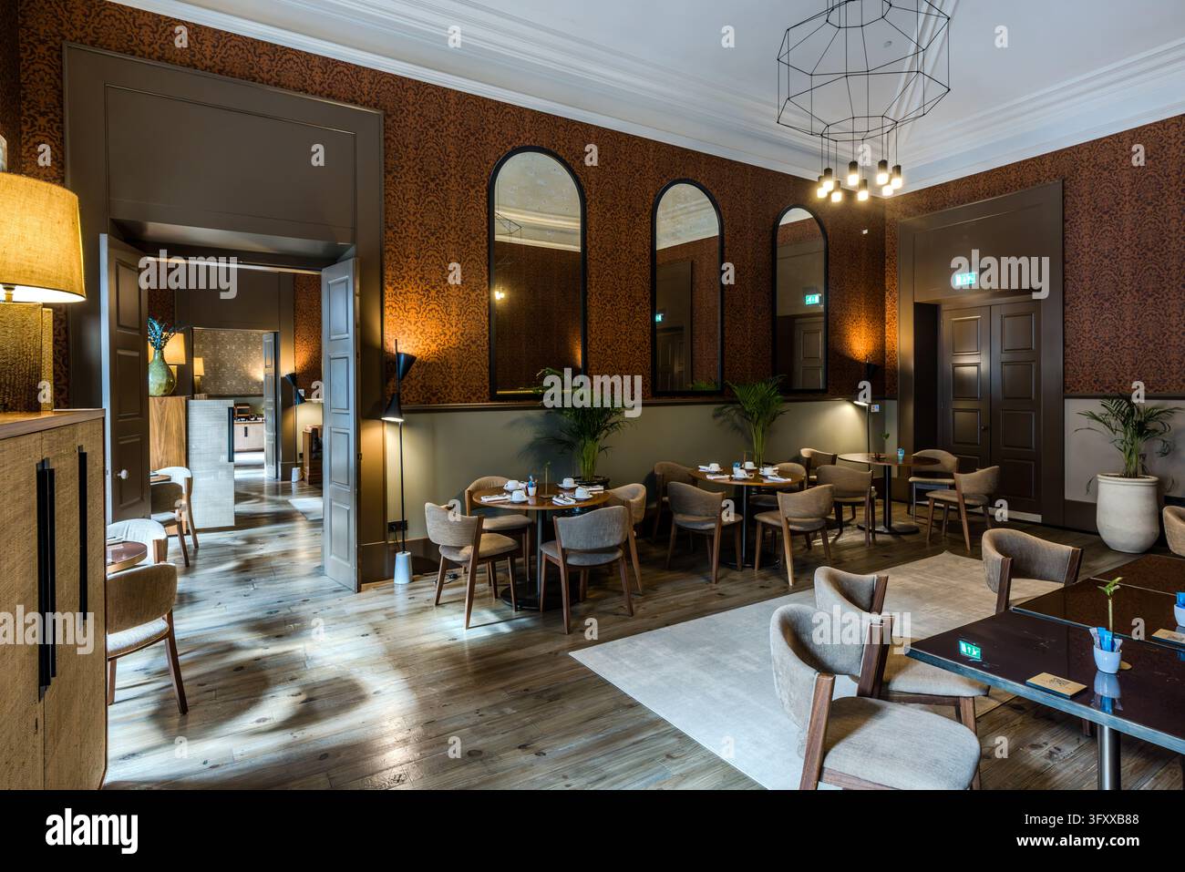 Ein Speisesaal mit gedeckten Tischen befindet sich möglicherweise im PortoBay Flores Hotel in Porto, Portugal. Braune gemusterte Tapeten bedecken die Wände, und drei gewölbte Spiegel mit schwarzen Rahmen hängen nebeneinander. Eine Leuchte aus dünnen Metallstäben hängt von der Decke. Stühle mit grauer Polsterung stehen an runden und rechteckigen Tischen auf einem Holzboden. Topfpflanzen stehen entlang der Wände. Das Hotel nutzt einen Palast aus dem 16. Jahrhundert. Stockfoto