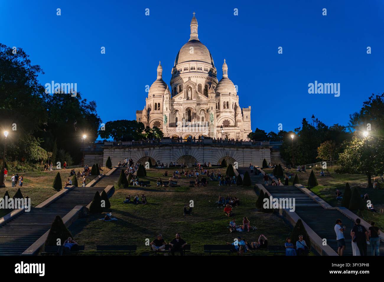 PARIS, FRANKREICH - 11. AUGUST 2026: Sacré-Cœur-Basilika bei Dämmerung beleuchtet mit Touristen auf den Stufen im Stadtteil Montmartre in Paris, Frankreich. Stockfoto