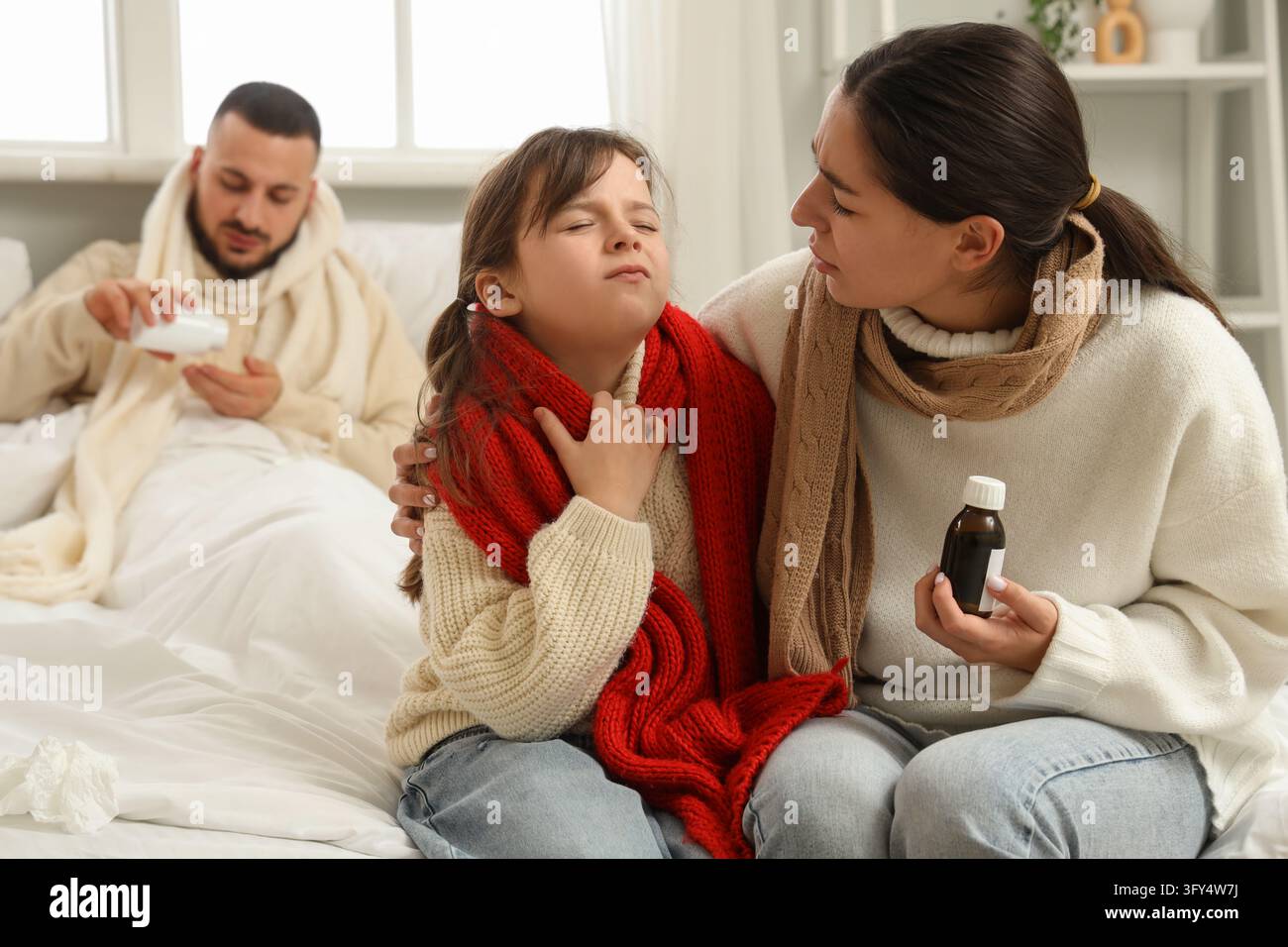 Familien-Gesundheitsversorgung und Grippesaison, Mutter kümmert sich um ihre kranke Tochter mit Halsschmerzen, während der Vater mit Erkältungssymptomen im Bett ruht, zu Hause im Schlafzimmer Stockfoto