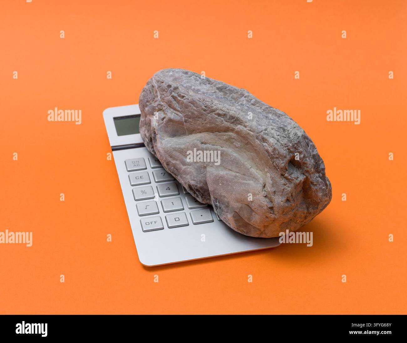 Schwerer grauer Stein, der einen silbernen Taschenrechner auf einem orangefarbenen Hintergrund niederdrückt, symbolisiert starken finanziellen Druck, schwere Steuerlast, wirtschaftliche Inflation Stockfoto