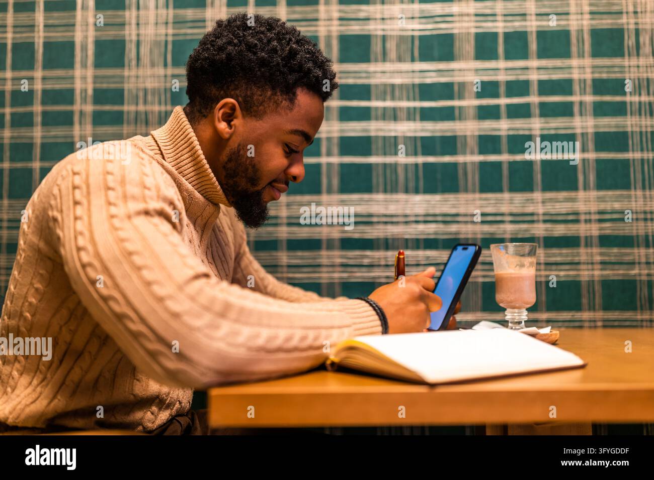 Junger Mann sitzt an einem Cafétisch, überprüft sein Smartphone und macht Notizen in einem offenen Notizbuch. Stockfoto