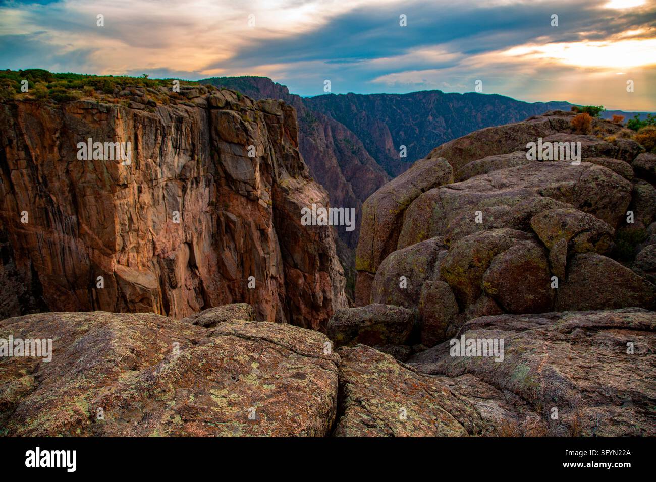 Ein malerisches Panorama der steilen, dunklen Felswände und der tiefen Schlucht des Black Canyon des Gunnison-Nationalparks bei Sonnenuntergang in Colorado, USA. Die Sonne wirft ein warmes, goldenes Licht auf die zerklüfteten Felswände, dramatische geologische Formationen und steile Abgründe, die im Laufe der Zeit vom Gunnison River geformt wurden. Die umliegende Landschaft zeigt spärliche Vegetation, die sich an die senkrechten Felswände klammert, und fängt die dramatische natürliche Schönheit und die raue Wildnis des amerikanischen Westens unter einem sanften Dämmerhimmel ein. Eine optimierte Version eines Bildes des US National Park Service. Credit: NPS / V Stauffenberg Stockfoto