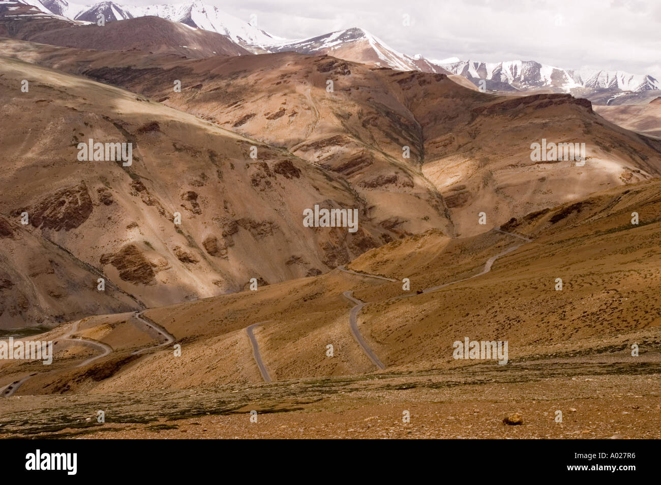 Kurvige Straße durch rötliche Felsformationen am Taglangla Pass am Manali-Leh Highway in Ladakh, Indien. Schneebedeckte Himalaya-Berge dahinter. Stockfoto