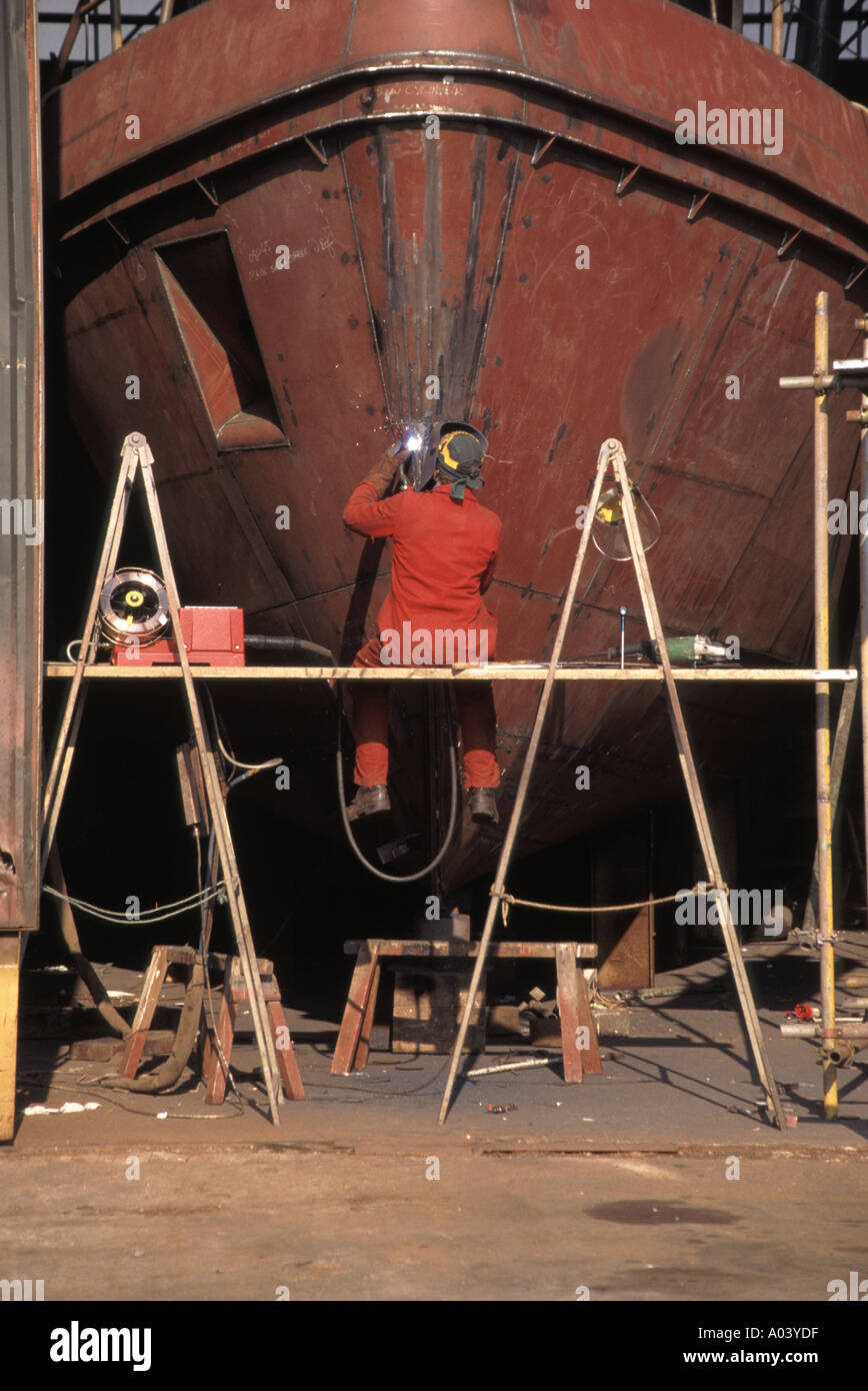 Ansicht der Werft Werkstatt & Rückansicht des Arbeiters auf Böcken sitzen bei der Arbeit an Schweißreparaturen Stahl Bug des Bootes in Werft Bristol England Großbritannien Stockfoto