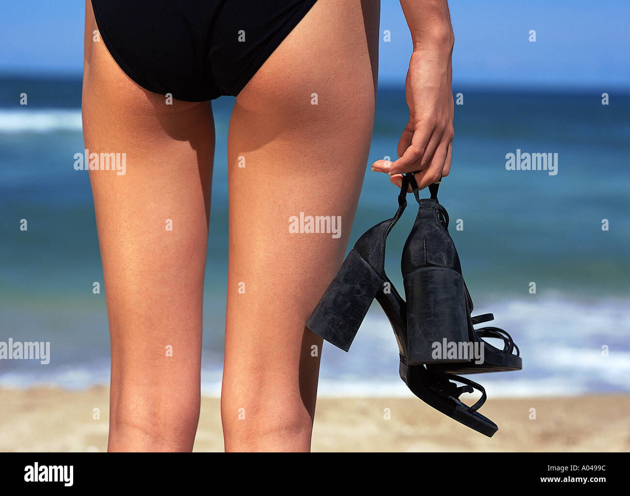 Detail-Aufnahme aus der unteren Hälfte des Körpers einer Frau stehend an einem Strand mit ein paar schwarze Schuhe in einer hand Stockfoto