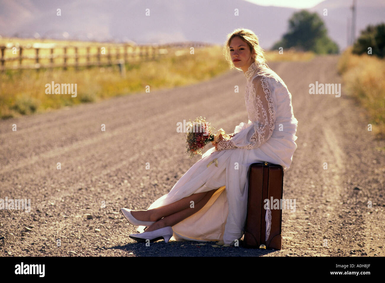 Junge Braut sitzt auf einem Koffer an der Seite einer Straße Stockfoto