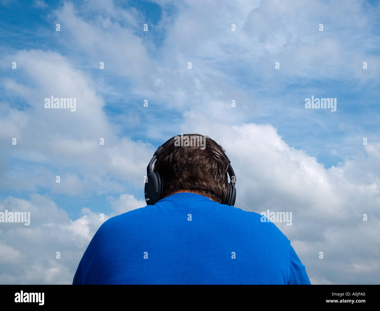 Großer Mann mit blauem Hemd anhören von Musik auf seine große Kopfhörer mit blauer Himmel mit Wolken im Hintergrund Stockfoto