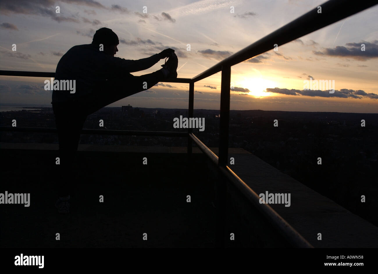Ein Läufer erstreckt sich auf einem Geländer mit einem Sonnenuntergang hinter ihm Stockfoto