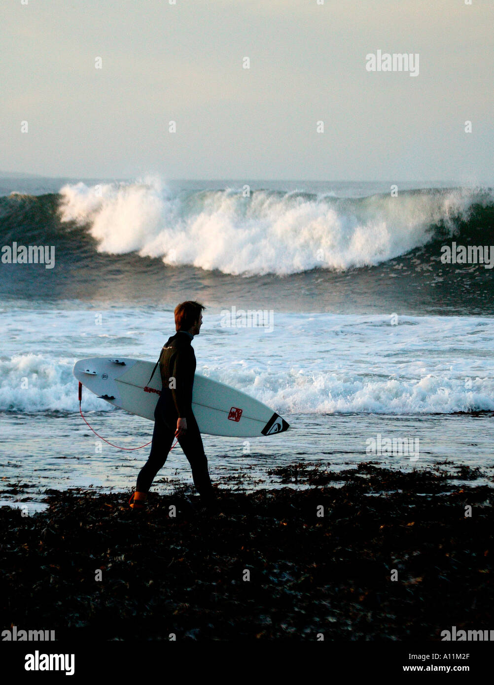 Einsamer Surfer fast in der Silhouette, die zu Fuß am Ufer mit Surfbrett und weißen Brandung im Hintergrund Stockfoto