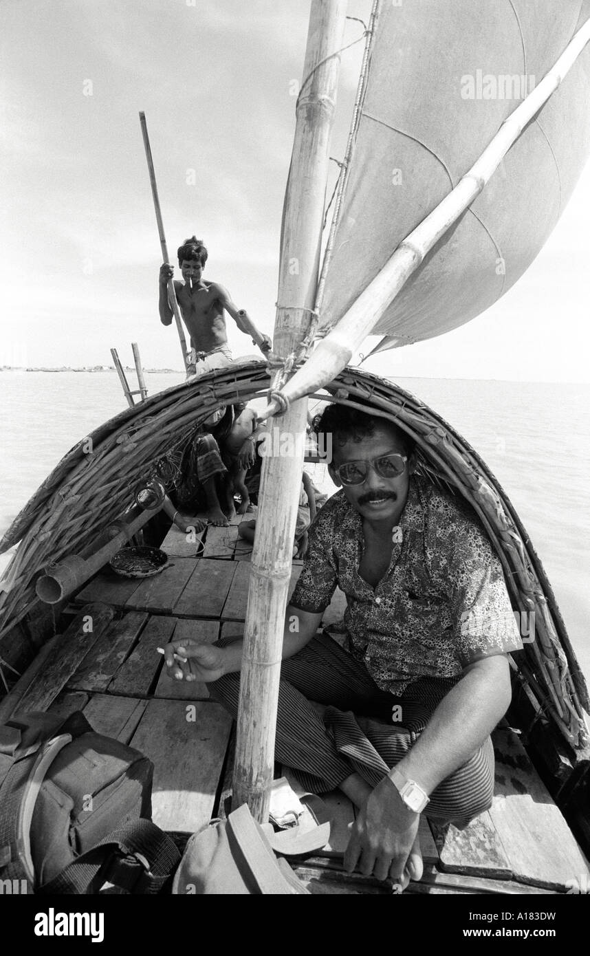 B/W eines Passagiers, der unter der Abdeckung auf einem kleinen Flussboot gerudert ist, rudert der Fährmann am hinteren Teil des Bootes an einem Zufluss des Ganges, Bangladesch Stockfoto
