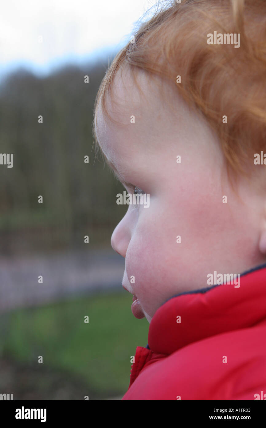 Baby mit roten Haaren tragen einen roten Körper wärmer außen Looking Weg von der Kamera zu spielen Stockfoto