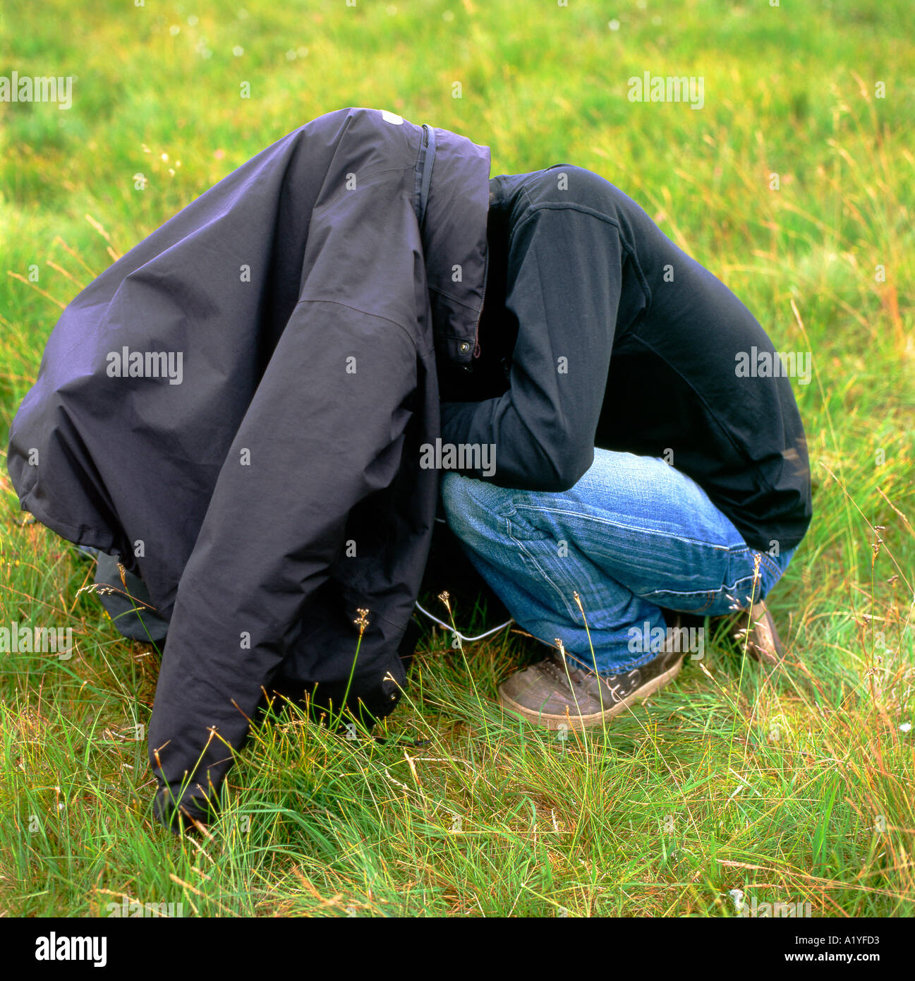 Mann mit Kopf unter seinem schwarzen Mantel Jacke hockend in einem Feld in Wales UK KATHY DEWITT Stockfoto