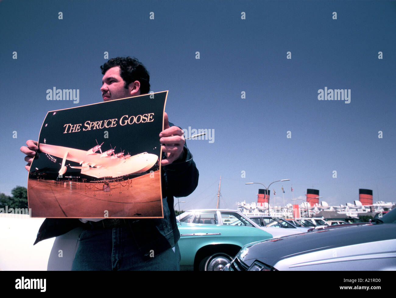 Mann hält auf einem Parkplatz mit den drei Trichtern der Queen Mary hinter Plakat von Howard Hughes Ebene "The Spruce Goose" Stockfoto
